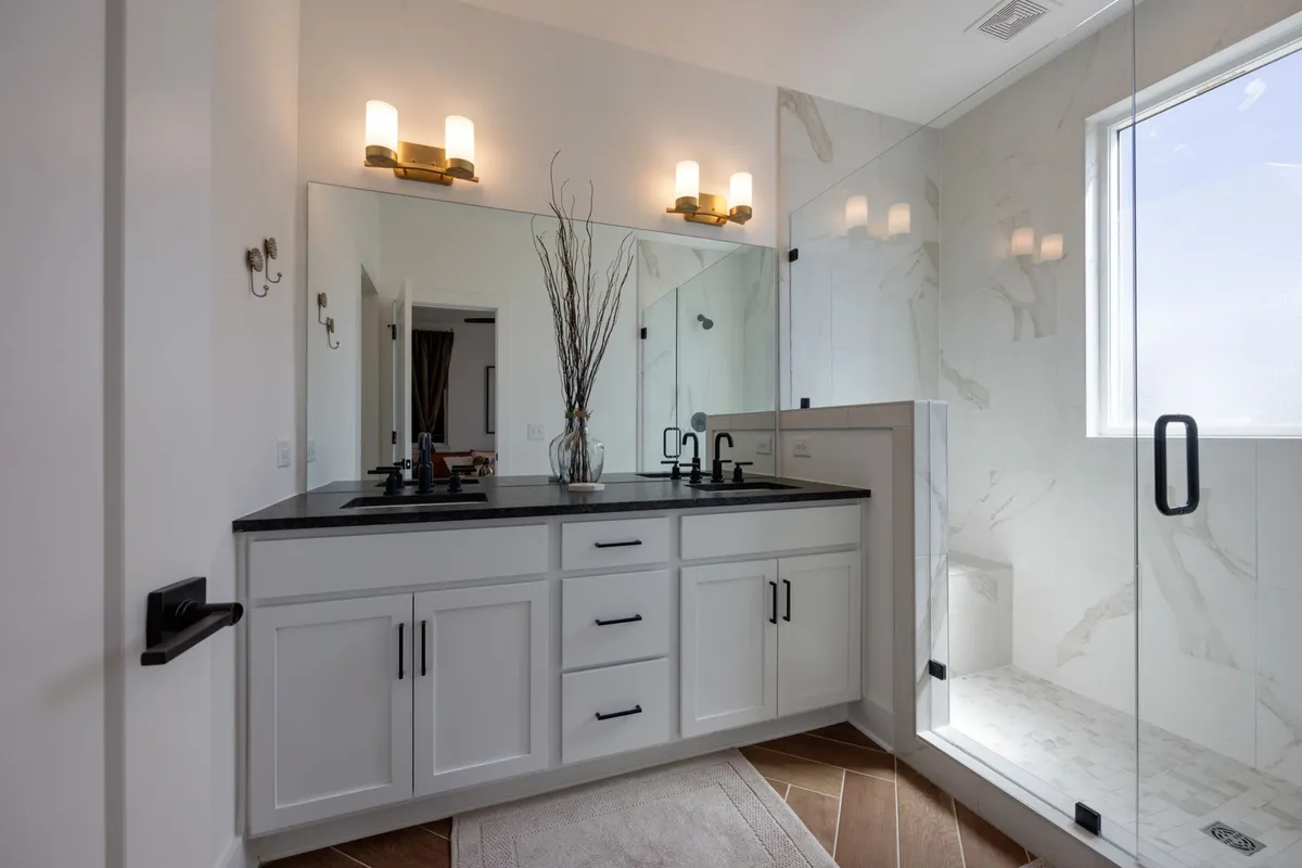 A modern, elegant bathroom with dual sinks, a large mirror, glass shower enclosure, and a decorative vase with branches. The space feels airy and sophisticated, ideal for self-care routines and privacy.