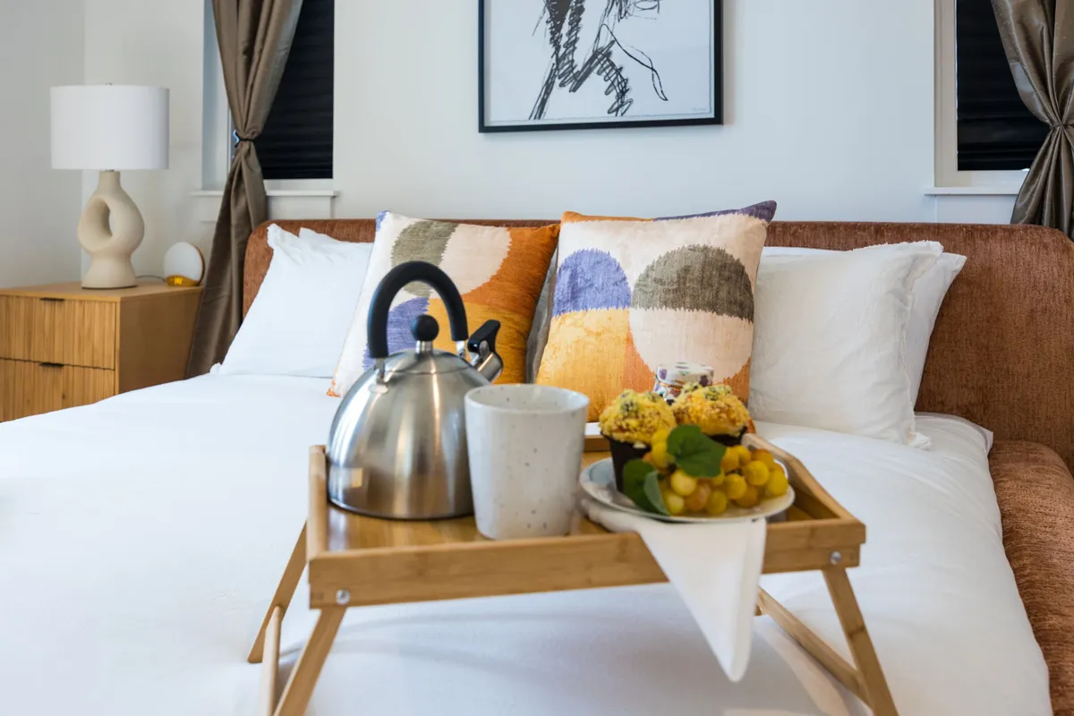 A welcoming bedroom with a bed topped with colorful pillows, a breakfast tray with a kettle and fresh fruit, and stylish artwork. The cozy lamp and window add to the inviting, personalized vibe.