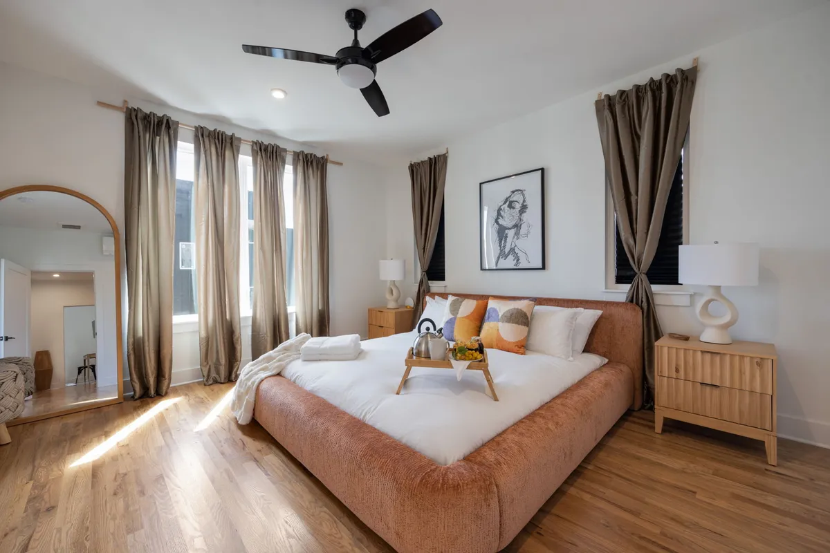 A serene bedroom featuring a large bed with soft, plush bedding, an art piece on the wall, and a mirrored dressing table. Natural light fills the space, creating a peaceful sanctuary ideal for relaxation and privacy.