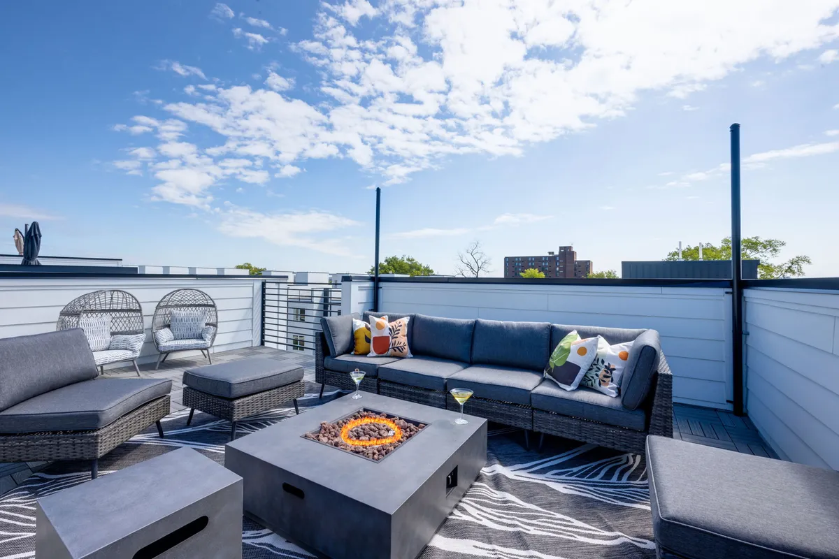 The image shows a stylish rooftop deck with a cozy outdoor seating area, a central fire pit, and decorative pillows, all set against the backdrop of Nashville's skyline. The scene invites relaxation, social gatherings, and enjoying the outdoors, with space for groups to unwind or entertain.