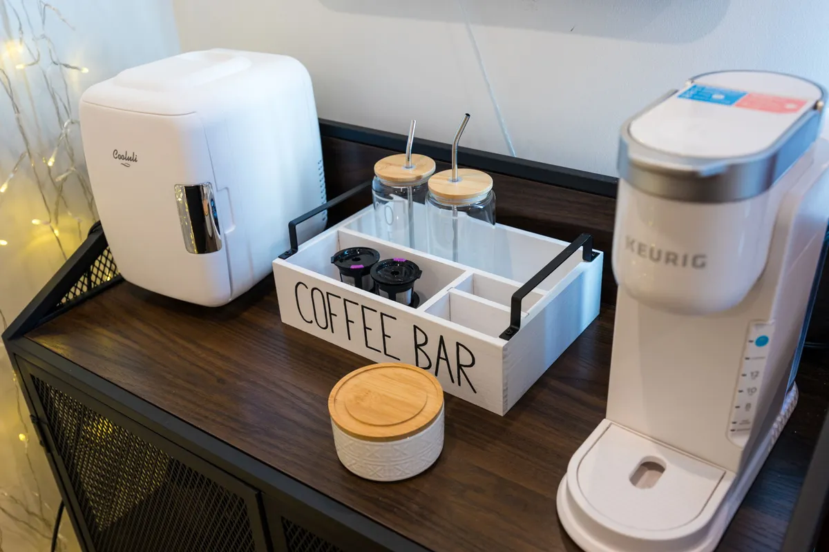 A dedicated coffee bar area with a Keurig coffee maker, mini refrigerator, and glass jars neatly arranged on modern shelving. The setup is cozy and modern, inviting guests to brew a fresh cup of coffee and enjoy a quiet morning or casual conversation.