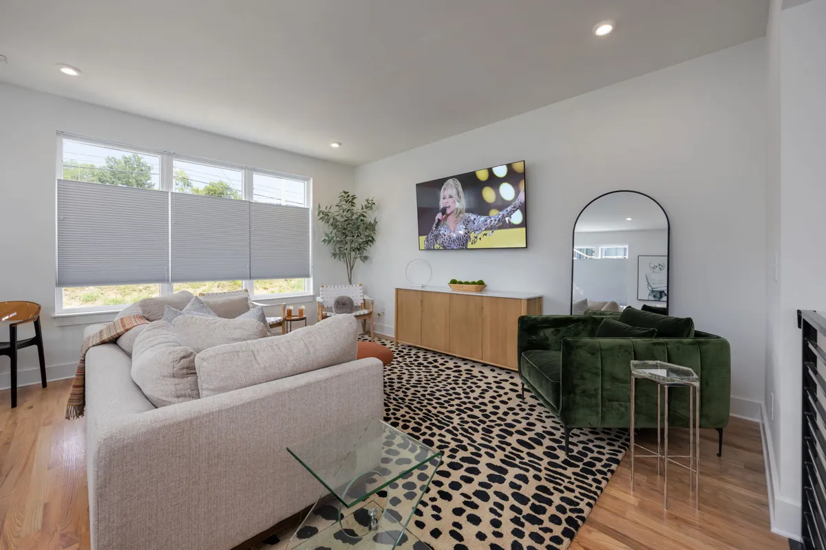 This image showcases a vibrant living room featuring a green velvet couch, a large television displaying a performer, a wooden sideboard, large windows letting in natural light, and a playful leopard print rug. The space is arranged to encourage lounging, socializing, and entertainment.